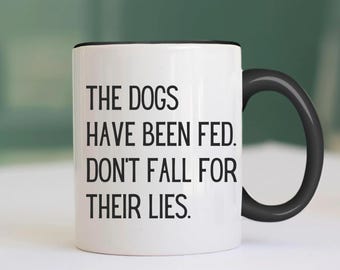 May include: White ceramic coffee mug with a black rim and handle. The mug has black text that reads "The dogs have been fed. Don't fall for their lies."
