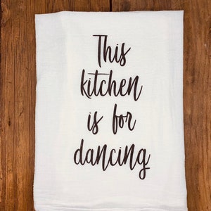 May include: White kitchen towel with black text that reads "This kitchen is for dancing."
