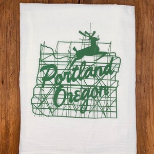 May include: White kitchen towel with a green outline of the state of Oregon, a stylized deer, and the text "Portland Oregon".