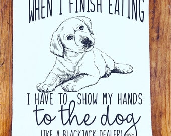 May include: A white tea towel with a black and white illustration of a dog. The text reads "WHEN I FINISH EATING I HAVE TO SHOW MY HANDS TO THE DOG LIKE A BLACKJACK DEALER!" with the copyright symbol and the letters "OVM".