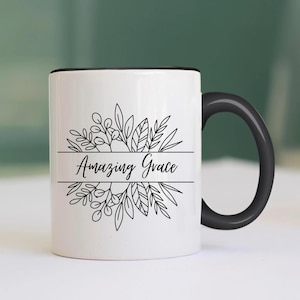 May include: White ceramic mug with a black rim and handle. The mug features a black line drawing of a floral wreath with the text "Amazing Grace" in the center.