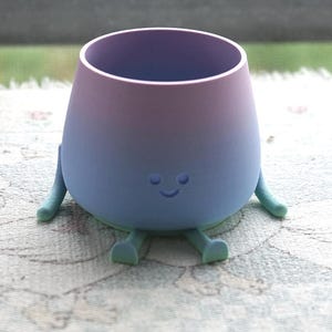 Könnte beinhalten: Eine skurrile, farbverlaufende Tasse mit einem lächelnden Gesicht, Armen und Beinen. Die Tasse geht von Lila in Blau über und erzeugt eine verspielte Ästhetik. Sie steht auf einem gemusterten, gefransten Tuch, was auf einen dekorativen oder neuartigen Artikel hindeutet.