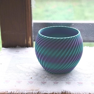 Puede incluir: Un jarrón esférico decorativo con un patrón arremolinado de color turquesa, morado y azul. El jarrón está sobre una superficie blanca cerca de una ventana. El jarrón tiene una abertura ancha y un exterior texturizado.