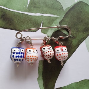 Puede incluir: Cuatro marcadores de punto en forma de búho con un pasador de seguridad plateado. Los búhos están pintados en azul y blanco, naranja y blanco, rojo y blanco, y rojo y blanco con detalles grises.