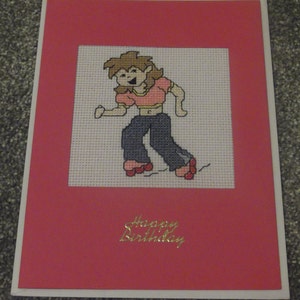 May include: A pink card with a cross-stitch image of a person roller skating. The card has the text "Happy Birthday" in gold lettering.