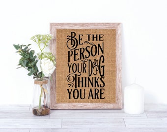 May include: A framed burlap sign with the text "Be the person your dog thinks you are" in black lettering. The sign is on a white surface with a white candle and a vase of white flowers.
