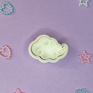 May include: A white plastic cookie cutter shaped like a cloud with a swirl design on a purple background. There are several small heart and star shapes made of pearls scattered around the cutter.