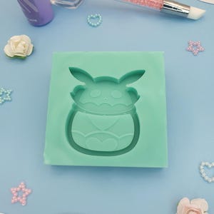 Puede incluir: Un molde de silicona verde claro con forma de personaje de dibujos animados. Una botella morada, una herramienta rosa y plateada y elementos decorativos como flores y corazones están dispersos alrededor del molde sobre una superficie azul claro.