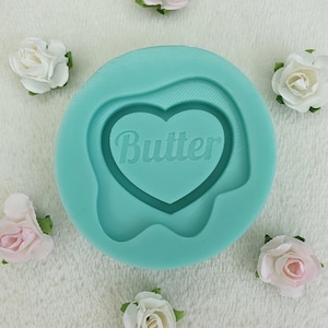 May include: A teal silicone mold with a heart-shaped design and the word "Butter" embossed inside. The mold is surrounded by small, artificial roses in shades of white and pink, arranged on a white, textured surface.