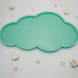 Puede incluir: Una bandeja de silicona en forma de nube de color verde claro con un borde elevado. La bandeja está rodeada de pequeñas flores blancas, estrellas rosas y corazones azules.