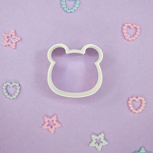 May include: White plastic cookie cutter shaped like a bear's head, surrounded by pastel pink, blue, and white pearl-like decorations on a purple background.