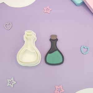 May include: A white plastic potion bottle cookie cutter next to a black and green potion bottle. The potion bottle has a brown cork. The background is purple with decorative stars and hearts.