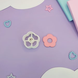 以下が含まれることがあります： 紫色の表面に、白いプラスチック製のフラワー型のクッキーカッターが2つあります。1つのカッターはシンプルなフラワー型で、もう1つは中央に切り抜きがあります。カッターの周りに、ピンクとブルーの真珠のような装飾が散らばっています。