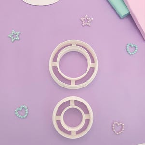 Puede incluir: Dos cortadores de galletas circulares blancos con un diseño en forma de cruz están centrados en una superficie lavanda. Dispersos hay adornos en forma de estrella y corazón con aspecto de perlas en colores pastel. Un recorte en forma de nube y bloques de colores pastel están en el fondo.