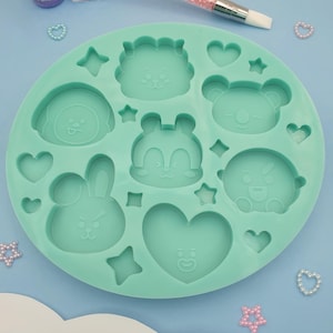 Può includere: Uno stampo rotondo in silicone verde acqua con varie forme di personaggi dei cartoni animati, tra cui un leone, un orso, un coniglio e un cuore. Lo stampo è circondato da piccoli abbellimenti a forma di stella e cuore. Un attrezzo con manico rosa e piccoli contenitori sullo sfondo.