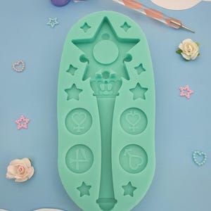 May include: A teal silicone mold with a star-shaped design, a wand with a crown, and four circular designs with symbols. The mold is on a light blue background.