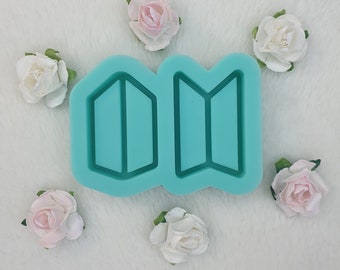 May include: A teal silicone mold in the shape of a stylized letter "M" with geometric segments. The mold is surrounded by small, artificial roses in shades of pink and white, arranged on a white, fluffy surface.
