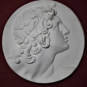 May include: A white plaster relief sculpture depicting a classical male profile. The artwork shows detailed curly hair and a prominent nose, set against a circular background. It is mounted with a small metal loop.