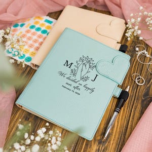 May include: A teal leather journal with a heart-shaped clasp and the text "We decided on happily ever after February 10, 2026." A beige journal and stickers are in the background. A pen and jewelry are also visible.