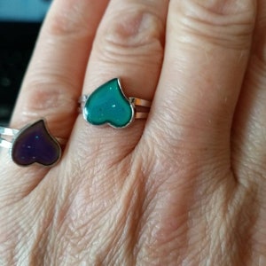 Può includere: Due anelli d'argento con pietre a forma di cuore. L'anello a sinistra ha una pietra viola, e l'anello a destra ha una pietra verde.