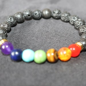 Könnte beinhalten: Armband mit dunklen, grauen Lavasteinperlen und sieben farbigen Chakra-Perlen. Die Chakra-Perlen sind in Regenbogenfarben angeordnet, von Violett bis Rot. Das Armband soll das Gleichgewicht fördern.