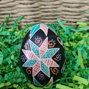 May include: A black, decorated egg with a starburst design in shades of pink, teal, and orange. The egg is surrounded by green paper grass, with a woven basket in the background. The egg is likely for Easter.