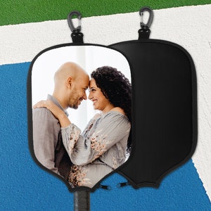 May include: Black pickleball paddle cover featuring a photograph of a couple. The cover includes a black carabiner clip and a zip closure. The paddle handle is visible at the base. The background is a blue and white court.