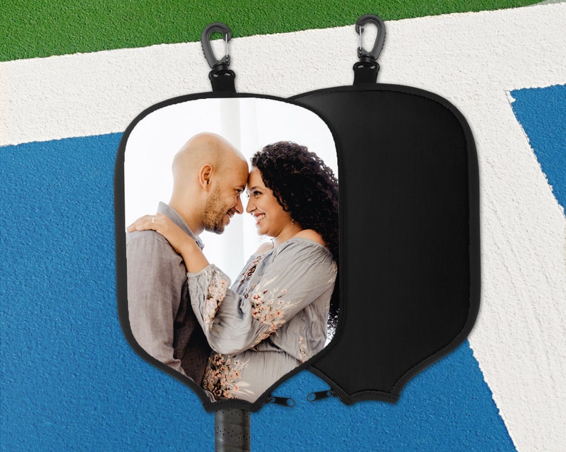 May include: Black pickleball paddle cover featuring a photograph of a couple. The cover includes a black carabiner clip and a zip closure. The paddle handle is visible at the base. The background is a blue and white court.