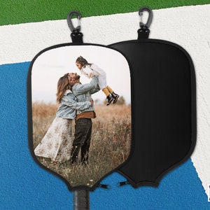 May include: A black paddleball paddle cover with a carabiner clip. One side features a family photo of a couple and a child. The other side is plain black. The paddle handle has a black grip.