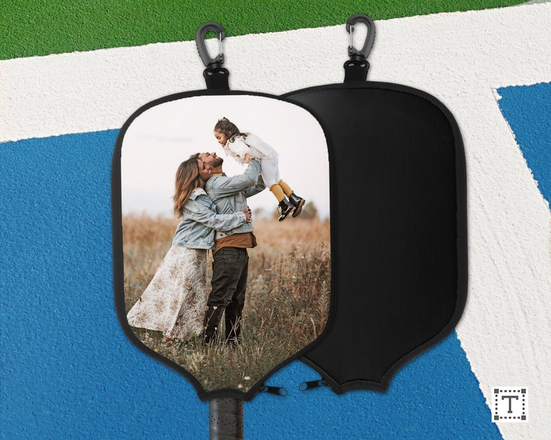 May include: A black paddleball paddle cover with a carabiner clip. One side features a family photo of a couple and a child. The other side is plain black. The paddle handle has a black grip.