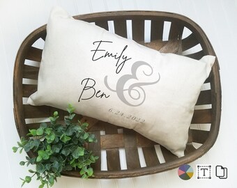 May include: A white pillow with the names "Emily & Ben" and the date "6.24.2022" printed in black. The pillow is in a wooden basket with green plants.
