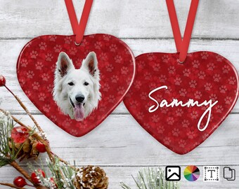 May include: Heart-shaped Christmas ornament with a red background covered in paw prints. One side features a white dog's face, the other side has the name "Sammy" in white script. A red ribbon is attached for hanging.