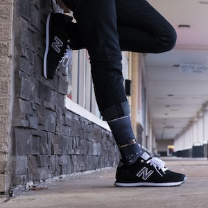 May include: A person wearing black jeans and black and white sneakers with a white 'N' logo. The person is standing with one leg bent and the other leg straight, leaning against a stone wall.