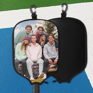 May include: Black pickleball paddle cover with a group photo. The cover has a black zip and a carabiner clip. The paddle handle is visible at the bottom. The background is a blue and white court.