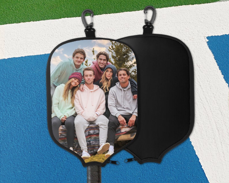 May include: Black pickleball paddle cover with a group photo. The cover has a black zip and a carabiner clip. The paddle handle is visible at the bottom. The background is a blue and white court.
