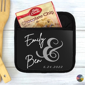 May include: Black pot holder with a pocket for a small item. The pot holder has the names "Emily & Ben" and the date "6.24.2022" printed on it in white.
