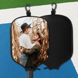 May include: Black pickleball paddle cover featuring a photograph of two people embracing in a cornfield. The cover has a black zip and a carabiner clip. The paddle handle is visible.