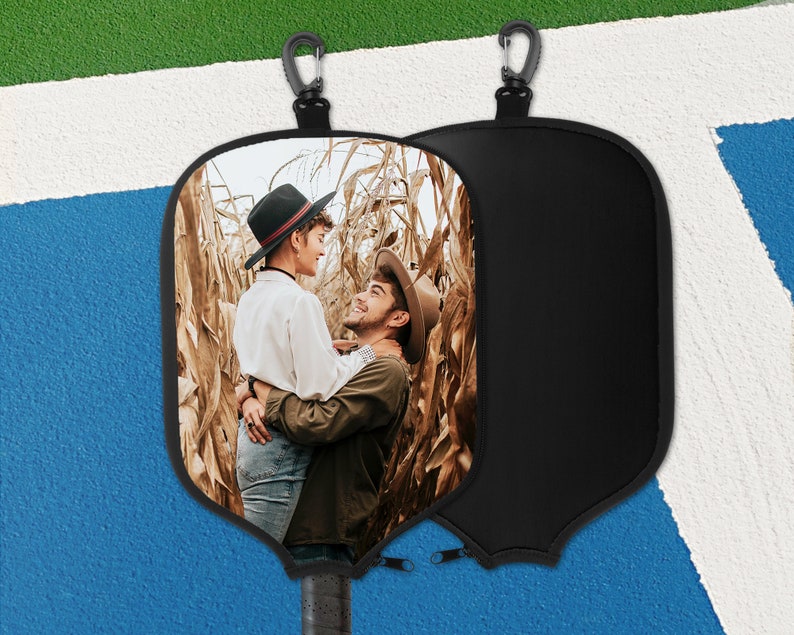 May include: Black pickleball paddle cover featuring a photograph of two people embracing in a cornfield. The cover has a black zip and a carabiner clip. The paddle handle is visible.