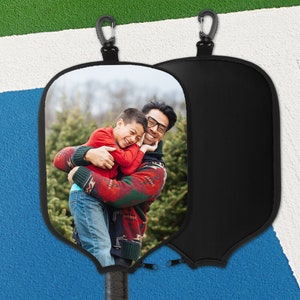 May include: Black pickleball paddle cover featuring a photograph of a father and child embracing. The cover includes a black zip and a carabiner clip. The paddle handle is visible at the base.