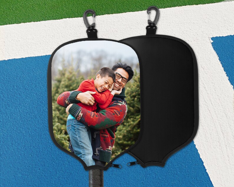 May include: Black pickleball paddle cover featuring a photograph of a father and child embracing. The cover includes a black zip and a carabiner clip. The paddle handle is visible at the base.