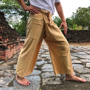 Può includere: Una persona indossa un paio di pantaloni marroni chiari con uno stile avvolgente. I pantaloni sono realizzati in un tessuto morbido e fluido e hanno una vestibilità rilassata. La persona è in piedi su un sentiero di pietra.