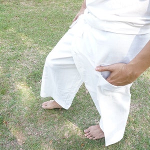May include: A pair of loose-fitting white wrap pants featuring a wide-leg silhouette, a high-waisted design with a tie closure, and a functional side pocket. The garment is constructed from a solid white fabric and reaches mid-calf length.