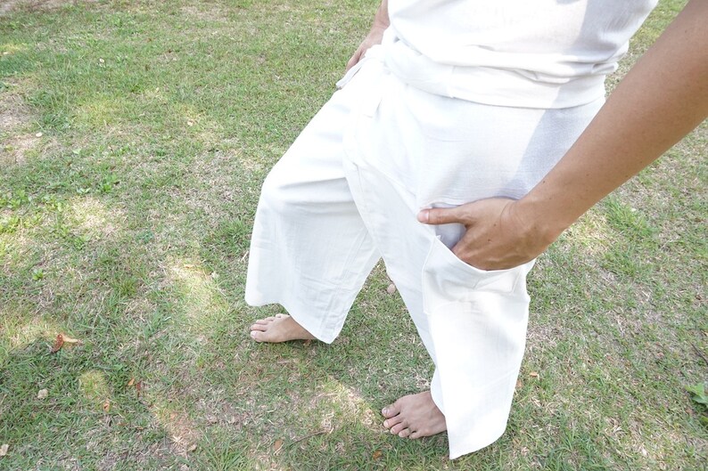 May include: A pair of loose-fitting white wrap pants featuring a wide-leg silhouette, a high-waisted design with a tie closure, and a functional side pocket. The garment is constructed from a solid white fabric and reaches mid-calf length.