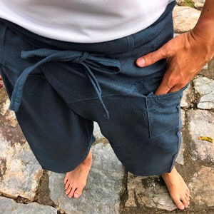 Pode incluir: Cal&ccedil;as envolventes azul escuro com fecho de la&ccedil;o na cintura e bolsos. As cal&ccedil;as s&atilde;o feitas de um tecido macio e s&atilde;o usadas com uma parte superior branca. A pessoa est&aacute; em p&eacute; sobre uma superf&iacute;cie de pedra.