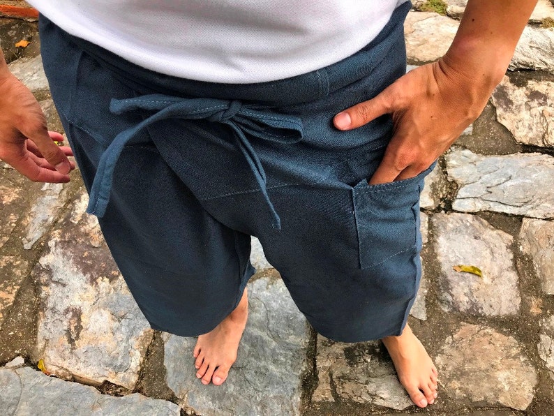 Pode incluir: Cal&ccedil;as envolventes azul escuro com fecho de la&ccedil;o na cintura e bolsos. As cal&ccedil;as s&atilde;o feitas de um tecido macio e s&atilde;o usadas com uma parte superior branca. A pessoa est&aacute; em p&eacute; sobre uma superf&iacute;cie de pedra.