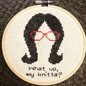 May include: Cross-stitch embroidery in a wooden hoop features a silhouette of a person with black hair and red glasses. The text "What up, my knitta?" is stitched below the figure. The background is white.