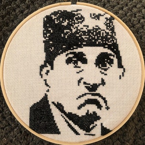 May include: A black and white cross-stitch portrait of a man in a wooden embroidery hoop. The image features a detailed depiction of the man's face, with black stitches forming his hair, eyes, mustache, and suit. The background is white.