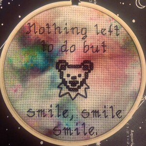 May include: Cross-stitch embroidery in a wooden hoop. The design features the text "Nothing left to do but smile, smile, smile" and a pixelated bear image, set against a colorful, tie-dye style background.