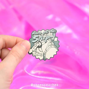 May include: A pin featuring a cartoon design of a harness and a penis, with the text "Money Get The Strap". The pin is in shades of grey, white, and black, with a light blue accent. The background is a bright pink.