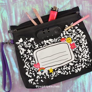 May include: A black zipped pouch featuring a white composition notebook pattern, a combination lock, and a purple wrist strap. The pouch includes a central white label graphic decorated with small stickers of a heart, rainbow, star, flower, lips, and smiley face.